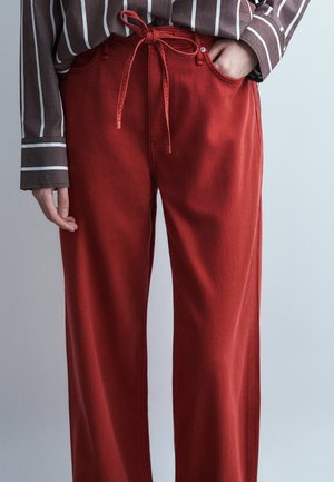 Személy laza, piros nadrágban, melynek dereka megkötött, barna csíkos hosszú ujjú ingben, egyik kezével a zsebében, a másik lazán pihen.