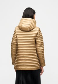 Γυναίκα με σκουρόχρωμα μαλλιά που φοράει ένα ταμπά, καπιτονέ μπουφάν με κουκούλα, στεκόμενη με την πλάτη προς την κάμερα μπροστά σε ένα απλό φόντο.