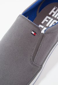 Scarpa slip-on grigia in tela con fodera interna blu. Presenta una piccola etichetta con logo in rosso, bianco e blu. Design semplice senza lacci.