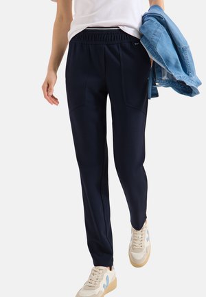 Persona che indossa pantaloni blu navy a sigaretta, sneakers bianche con dettagli blu, camicia bianca, che tiene una giacca di jeans azzurro chiaro piegata in una mano.