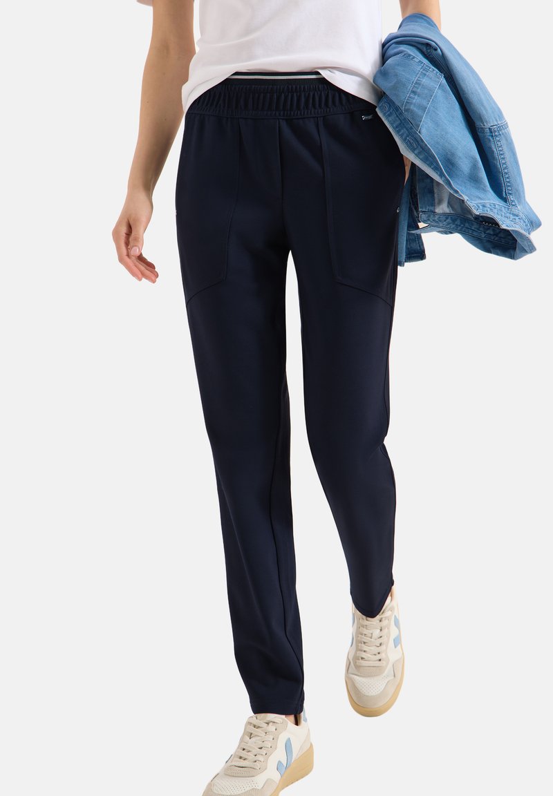 Personne portant un pantalon bleu marine fuselé, des baskets blanches avec des accents bleus, une chemise blanche, tenant une veste en denim bleu clair pliée dans une main.