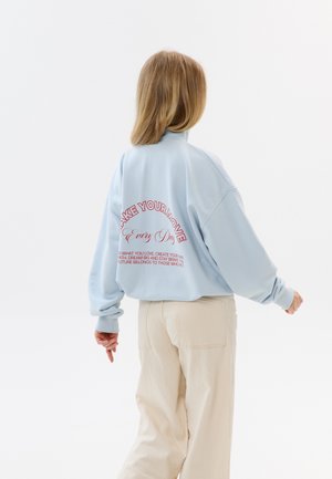 Osoba z blond włosami do ramion, ubrana w jasnoniebieską bluzę z czerwonym napisem i beżowe spodnie, zwrócona tyłem na jednolitym białym tle.