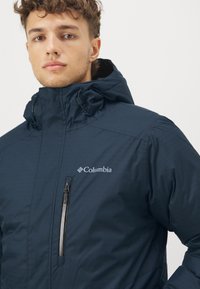 Marineblaue Columbia-Jacke mit hohem Kragen, verstellbarem Kapuze und einer mit einem silbernen Reißverschluss versehenen Brusttasche. Glattes, leichtes Material.