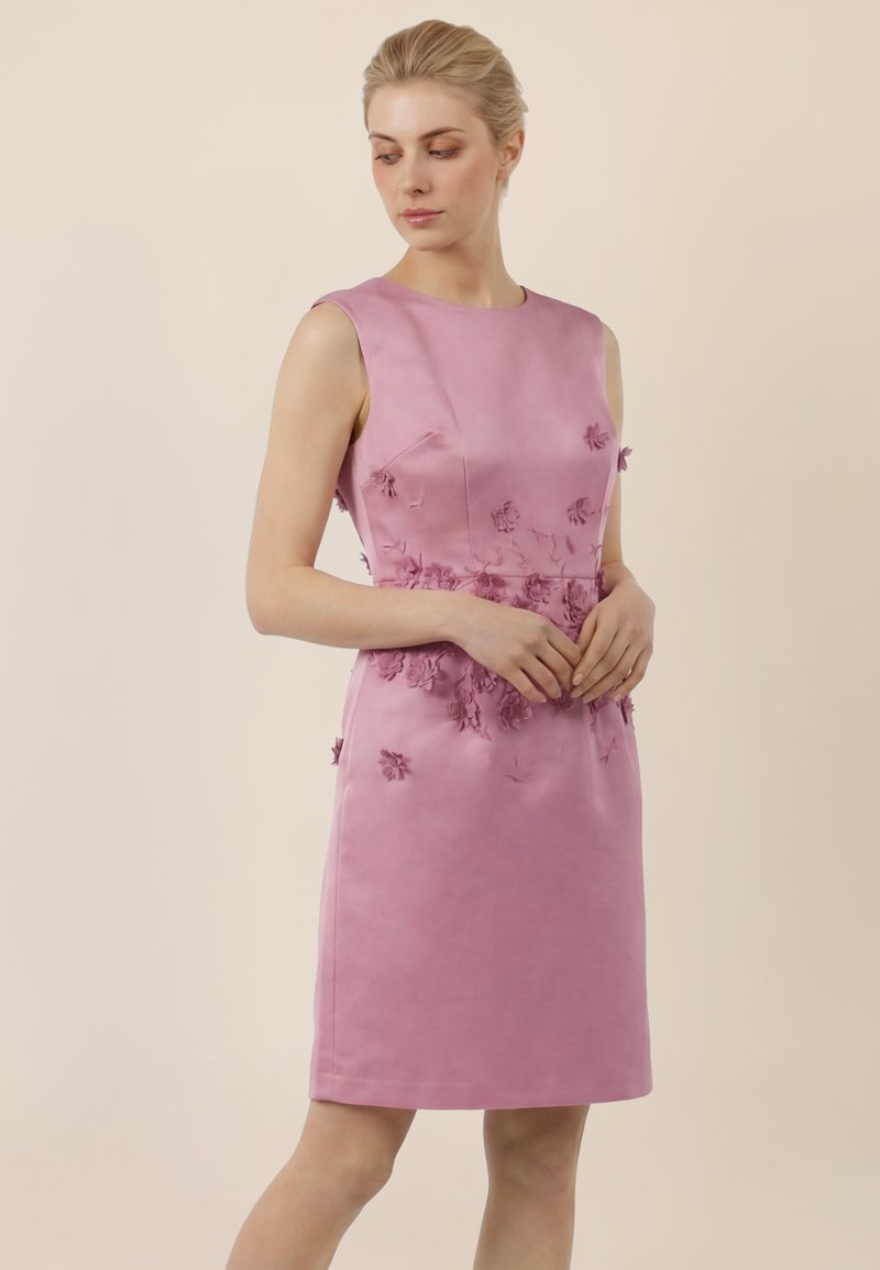 Femme portant une robe rose sans manches, longueur genou, ornée d'appliqués floraux 3D sur le corsage et la taille, debout devant un fond beige uni.