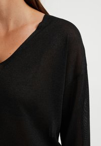 Top negro ligero de punto con un escote en V profundo, mangas largas y una textura translúcida. El dobladillo y el collar tienen un acabado ligeramente áspero.