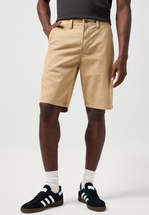 Pantaloni corti beige in cotone con taglio dritto, tasche laterali e chiusura a bottone, indossati con sneakers blu navy con strisce bianche e calze alla caviglia.