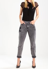Top negro con lunares y un lazo, combinado con jeans gris desgastados que tienen bordados de rosas y estrellas, y tacones altos negros.