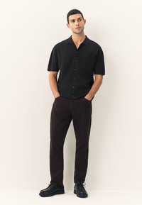 Svart kortärmad skjorta med knappar och en bröstficka, kombinerad med mörkbruna byxor och svarta skor, placerad mot en neutral bakgrund.