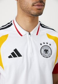 Hvit polo skjorte med glidelåskrage, med svarte, røde og gule aksenter. Inkluderer en svart Adidas-logo og et tysk fotballforbund merke.