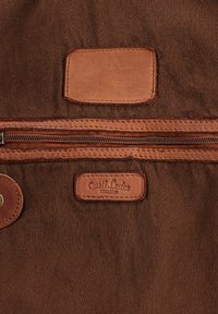 Brown fabric surface with horizontal zipper and two stitched leather patches, one embossed with "Gusti Leder studio" text.