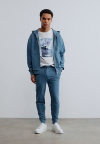 Νέος άνδρας όρθιος, φορώντας μπλε φούτερ με κουκούλα, ταιριαστό παντελόνι φόρμας, λευκό T-shirt με σχέδιο και λευκά αθλητικά παπούτσια, σε απλό φόντο.