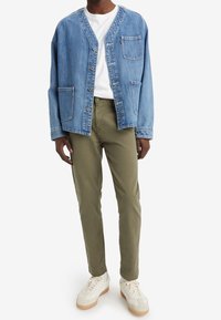 Cardigan in denim blu con scollatura a V, chiusura a bottoni e due tasche, abbinato a pantaloni verdi oliva e sneakers bianche.