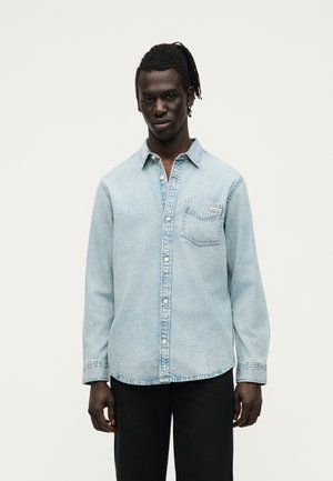 Mies, jolla on tumma iho ja dreadsit, pukeutuneena vaaleansiniseen farkkupaitaansa ja mustiin housuihin, seisoo yksinkertaisen valkoisen taustan edessä.