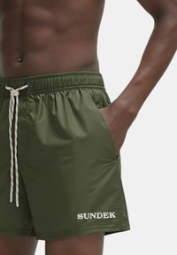 Bañadores de color verde oliva en material ligero, con cintura elástica, cierre de cordón, bolsillos laterales y logo "SUNDEK" en blanco.