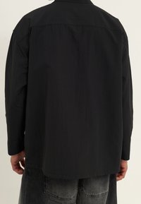 Black shirt with a structured design, featuring long sleeves, a collar, and a smooth texture. The back has a center seam and relaxed fit.