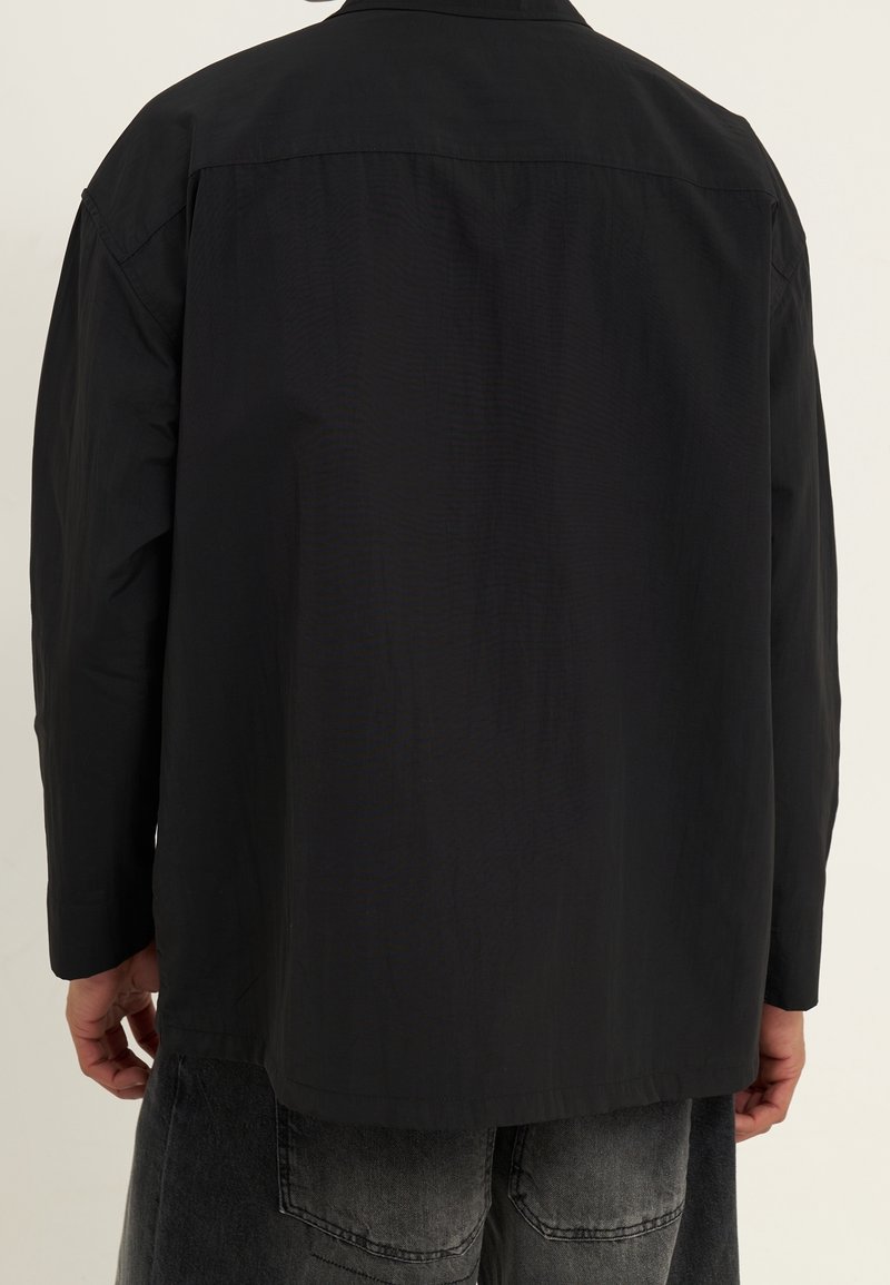 Black shirt with a structured design, featuring long sleeves, a collar, and a smooth texture. The back has a center seam and relaxed fit.