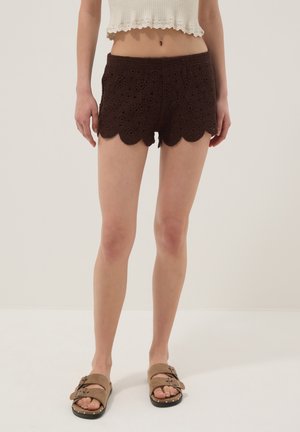 Mujer vistiendo pantalones cortos de ganchillo marrón oscuro, top de punto sin mangas beige y sandalias marrón claro con hebilla, de pie sobre fondo neutro.