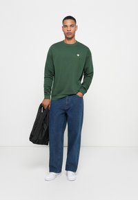 Felpa verde a girocollo, vestibilità rilassata. Jeans blu scuro, a vestibilità ampia. Porta una borsa tote nera, con texture. Sneakers bianche completano il look.