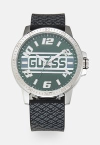 Quarzuhr mit einem Gehäuse aus silbernem Edelstahl und grünem Zifferblatt. Schwarzes Gummiband mit geometrischem Muster. Große Stundenmarkierungen.