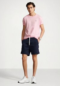 Rožinis medvilninis marškinėliai su apvalia iškirpte, derinami su tamsiai mėlynomis šortais su elastinga juosmens juosta ir raišteliais. Baltos sportinės bateliai.