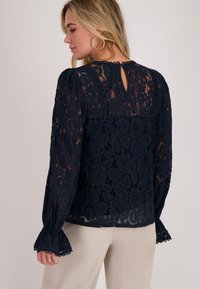 Marineblauwe kanten blouse met een bloemenpatroon, lange pofmouwen en een keyhole-detail op de rug. Geschulpte manchetten zorgen voor textuur en elegantie.