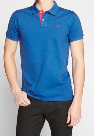 Mand iført blå poloshirt med rød knaplukning og sorte bukser, står med én hånd i lommen på hvid baggrund.