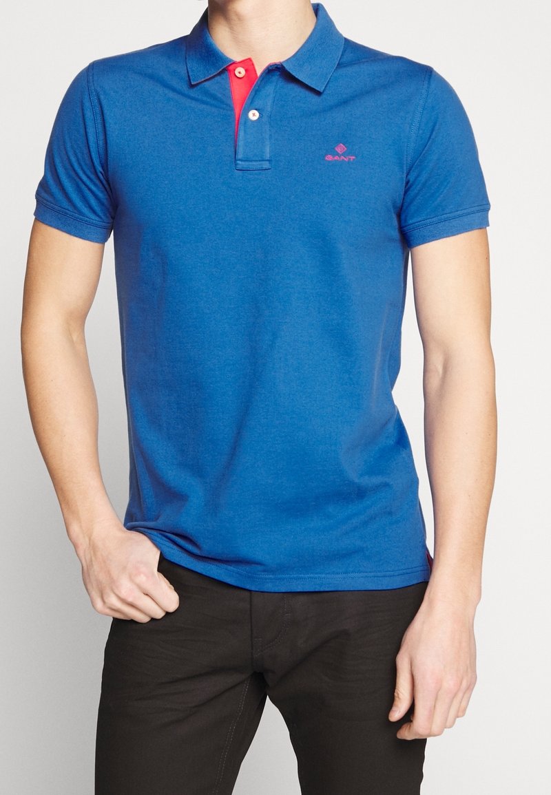 Homme portant un polo bleu avec une patte de boutonnage rouge et un pantalon noir, debout avec une main dans la poche sur fond blanc.