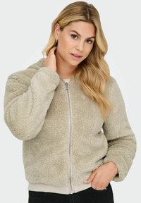 Fuzzy beige zip-up jacket with a hood, ribbed hem, and cuffs, featuring a soft texture and a relaxed fit. Model with long hair shows detail.