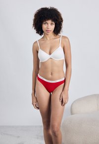 Λευκό σουτιέν με λεπτές τιράντες και κόκκινο στρινγκ με λευκή λογότυπη κορδέλα. Λείο υλικό, μινιμαλιστικός σχεδιασμός, περιγραμματική μορφή.