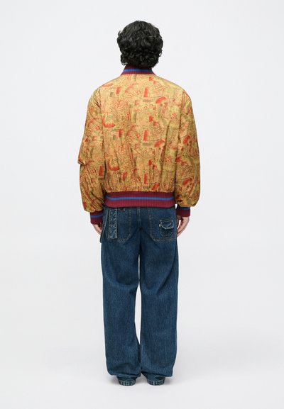 Zlatá bomber bunda s farebnou potlačou, rebrovaným golierom a manžetami, v kombinácii s voľne strihanými modrými džínsami s zadnými vreckami.