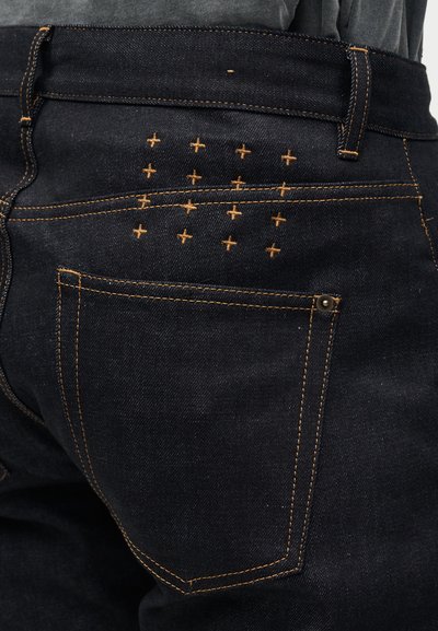 Σκούρο μπλε τζιν παντελόνι με πορτοκαλί ραφές στην πίσω τσέπη και σταυροειδές κέντημα πάνω από την τσέπη.