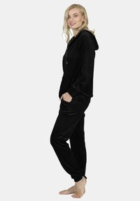 Black velour tracksuit with a zip-up hoodie and front pockets, featuring a smooth texture and tapered pants, worn barefoot.
