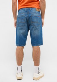 Szorty dżinsowe w średnim niebieskim kolorze, o klasycznym kroju, z widocznym szwem, dwoma tylnymi kieszeniami z naszywką marki oraz postrzępionym dołem.