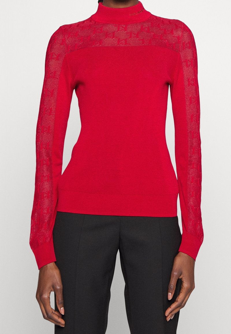 Maglione rosso a maglia con collo alto e maniche trasparenti a motivo. Design aderente, texture liscia e polsini a costine. Abbinato a pantaloni neri.