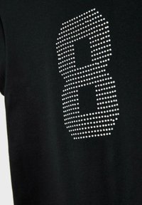 Crna majica s velikim brojem "8" od srebrnih rhinestona, koji stvaraju teksturirani uzorak. Glatka tkanina s uprivatom kroju.