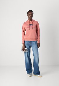 Felpa rosa con tasca frontale, caratterizzata da un logo e un anno. Abbinata a jeans chiari a zampa d'elefante e sneakers argentate, mentre tiene una pochette argentata.