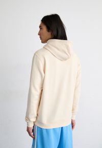 Felpa leggera beige con una texture morbida, dotata di cappuccio con cordoncino e polsini a coste, abbinata a pantaloni sportivi blu.