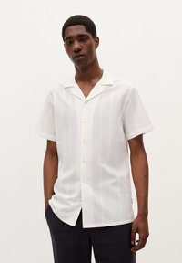 Camisa de manga corta blanca con cierre frontal de botones, que presenta rayas verticales y botones amarillos. Tejido suave, corte relajado, cuello estándar.