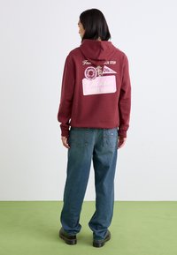 Burgundy hooded sweatshirt with a graphic print featuring donuts and text. Paired with loose-fitting blue denim pants and black shoes.