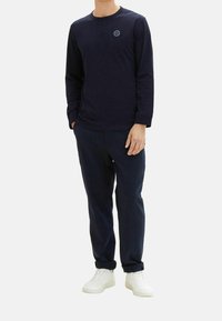 Marinblå långärmad t-shirt med rund halsringning och liten logotyp på bröstet, ihopparad med matchande marinblå byxor och vita sneakers.