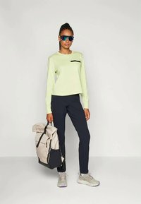 Fényzöld, hosszú ujjú felső fekete mellzsebbel, navy kék nadrággal párosítva. A modell egy bézs és navy kék hátizsákot tart. Szürke sportcipő, lila részletekkel.