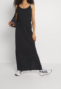 Robe maxi noire à fines bretelles, taille cintrée et fente latérale. Assortie à des baskets blanches et noires ainsi qu'un petit sac noir.