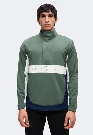 Homme portant un pull Nike Golf vert, blanc et bleu marine avec un col montant et un logo sur la poitrine, debout devant un fond clair uni.