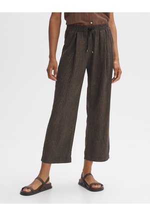 Mujer con pantalones sueltos marrones con estampado y cintura elástica, y sandalias negras de punta abierta, de pie frente a un fondo liso.