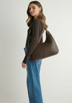 Kobieta ubrana w ciemnobrązową bluzkę z długim rękawem i niebieskie dżinsy niesie dużą ciemnobrązową skórzaną torbę na ramię, stojąc na jasnym tle.