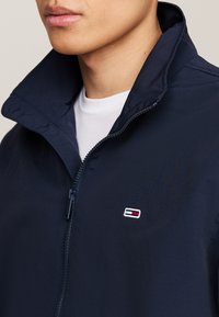 Mornarska zip-up jakna z visokim ovratnikom, ki ima majhen vezen logotip na prsih. Gladka tekstura, lahka tkanina in brez vidnih pripomočkov.