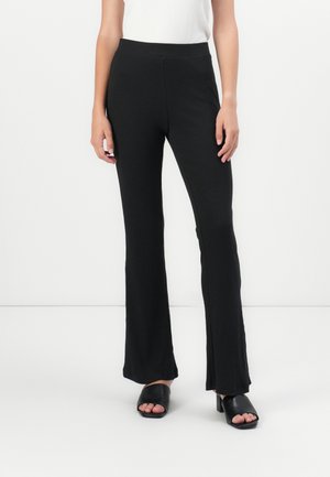 Pantalons noirs à côtes évasés avec une taille haute, texture lisse et fentes latérales, associés à des chaussures noires à talons ouverts.