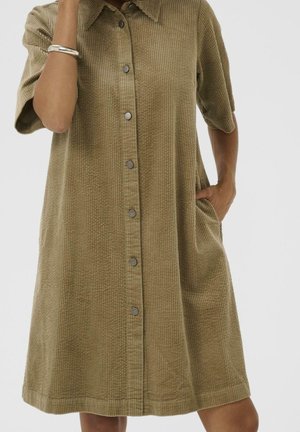 Femme portant une robe en velours côtelé courte aux genoux, manches courtes, boutonnée sur le devant, avec col et poches latérales, debout devant un fond uni.