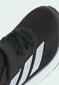 Zapatilla deportiva negra con parte superior de malla texturizada, rayas blancas en contraste y un cuello acolchado para mayor comodidad. Punta redondeada y suela de goma.