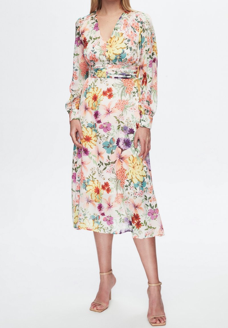 Femme portant une robe midi à manches longues, col en V, avec un motif floral coloré et des sandales beiges à talons hauts avec bride à la cheville.